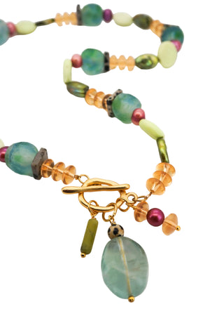 A beaded necklace with a gold toggle clasp, featuring translucent light green beads, textured green stone beads, amber-orange faceted beads, and a large hanging oval light green stone charm, displayed on a white background.